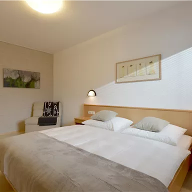 A modern bedroom with two single beds, a comfortable armchair, and soft lighting. The walls are light and minimally decorated.