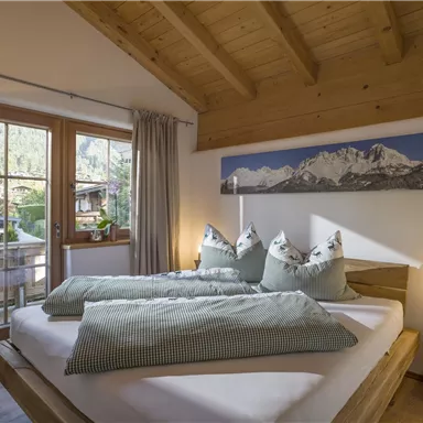 Ein gemütliches Schlafzimmer mit einem großen Fenster und Holzdetails. Die Betten sind mit grünen Kissen und Decken dekoriert.