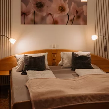 A cozy bedroom with a large bed and soft pillows. On the wall hangs a picture of beautiful, pink flowers, and a simple lamp provides gentle light.