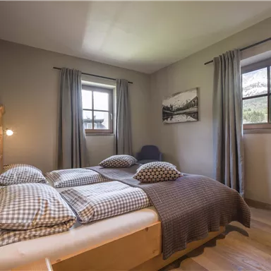 A cozy bedroom with a large bed and checkered pillows. Windows offer a view of nature, while curtains gently filter the light.