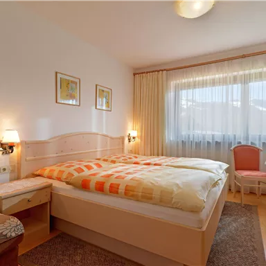 A cozy bedroom with a large double bed and bright curtains. A small table and chairs complement the space, which appears friendly and inviting.