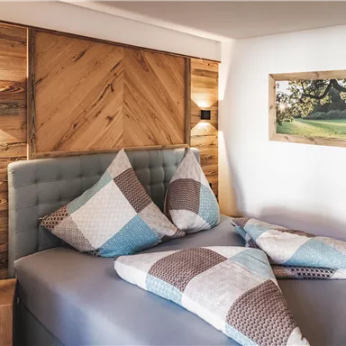 Ein modernes Schlafzimmer mit einem gemütlichen Bett und bunten Kissen. An der Wand befindet sich ein Holzpaneel und ein landschaftliches Bild.