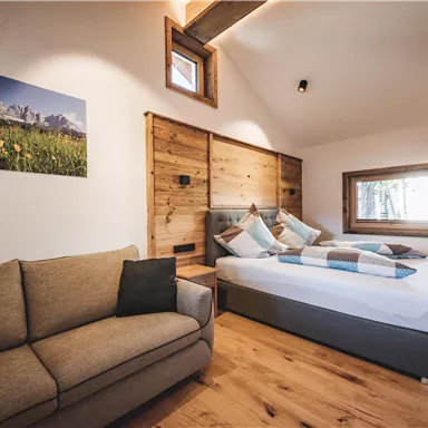 Ein modernes Schlafzimmer mit einem Doppelbett und einem gemütlichen Sofa. Holzverkleidungen und große Fenster sorgen für eine helle, einladende Atmosphäre.