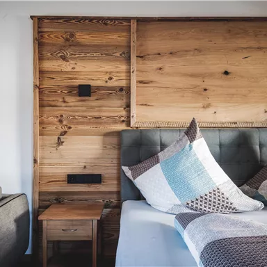 Ein gemütliches Schlafzimmer mit einem Holzpaneelkopfteil und dekorativen Kissen. Links befindet sich ein graues Sofa und ein Nachttisch.