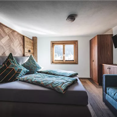 Ein modernes Schlafzimmer mit einem gemütlichen Bett, bunten Kissen und einem kleinen Sofa. Ein Fenster mit Blick nach draußen sorgt für Tageslicht.