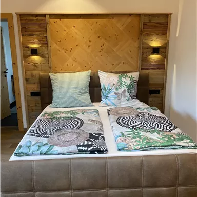 Ein komfortables Doppelbett mit farbenfrohen Decken und Kissen. Der Kopfbereich ist aus Holz und wird von modernen Wandlampen beleuchtet.