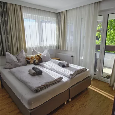 A bright bedroom with a double bed and two pillows. Large windows provide plenty of daylight and offer access to a balcony.