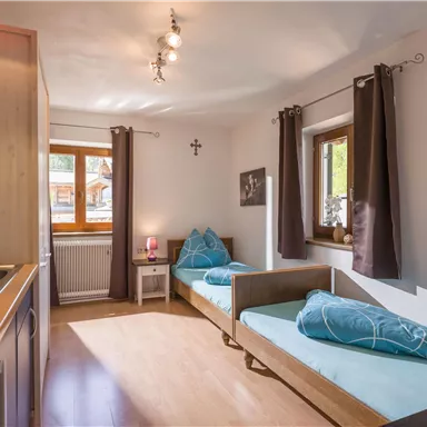 A cozy room with two single beds and blue cushions. The windows provide daylight, and the kitchen is conveniently integrated.