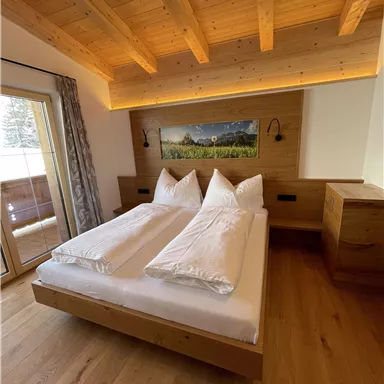 A cozy bedroom with a wooden bed and two large pillows. The room has large windows and a warm wood look.