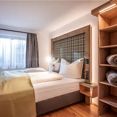 Ein modernes Schlafzimmer mit einem Doppelbett und Holzregalen. Helle Gardinen und eine warmen Farbgestaltung schaffen eine gemütliche Atmosphäre.