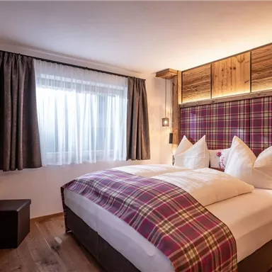 Ein gemütliches Schlafzimmer mit einem großen Bett und karierten Kissen. Das Zimmer hat Holzverkleidung und ein Fenster mit Vorhängen, das natürliches Licht hereinlässt.