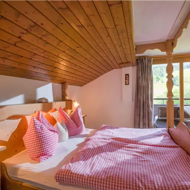 A cozy bedroom with wooden paneling and red-and-white checkered pillows. There is a balcony with a view of nature.