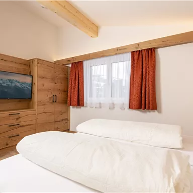 A cozy bedroom with two beds and warm wooden furniture. Large windows and decorative curtains create an inviting atmosphere.