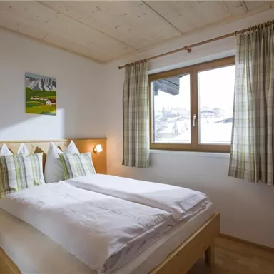 Ein gemütliches Schlafzimmer mit einem Doppelbett und Holzregalen. Große Fenster sorgen für viel Tageslicht und Ausblick.