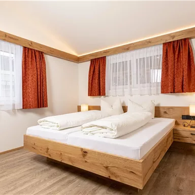 A bright bedroom with a large double bed and wood details. The windows are decorated with white curtains and red drapes.