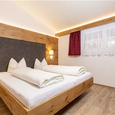 A cozy bedroom with a wooden double bed. The walls are light, and the room has large windows with red curtains.