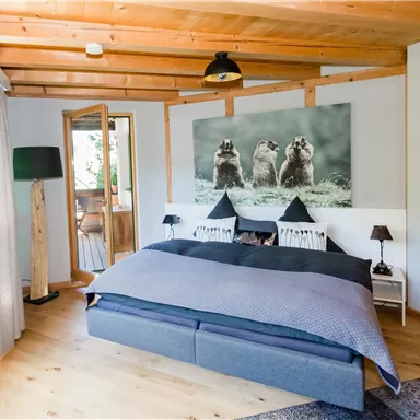 Ein gemütliches Schlafzimmer mit einem großen Bett und warmem Holzdekor. An der Wand hängt ein Bild von drei Tieren, und es gibt viel natürliches Licht durch die Fenster.