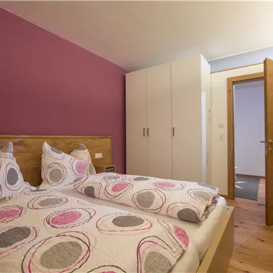 Ein gemütliches Schlafzimmer mit einem Doppelbett und bunten Kissen. Die Wände sind in einem sanften Lila gestrichen, und die Tür ist offen, um einen Blick in den Flur zu zeigen.