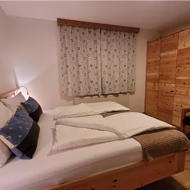 A cozy bedroom with a large bed and wooden-colored furniture. The windows are dressed with curtains, and there is a small seating area in the room.