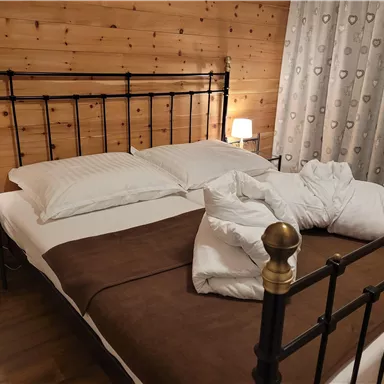 A cozy bedroom with a metal bed and white pillows. The walls have wooden panels and the windows are decorated with curtains.