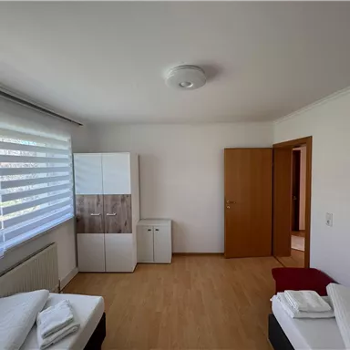 A simple and bright bedroom with two beds and wooden floor. There is a wardrobe and another room in the background.