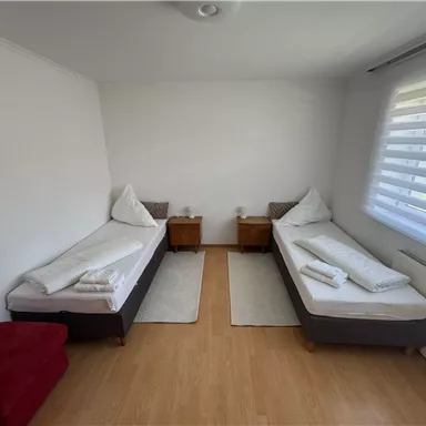 A bright room with two single beds and bedside tables. The walls are white and there are large windows with blinds.