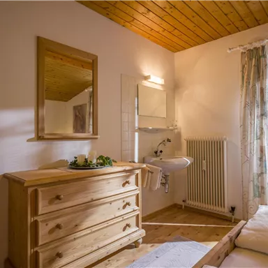 Ein gemütliches Schlafzimmer mit Holzfußboden und einem großen Fenster. Die Einrichtung umfasst eine Holzkommode und einen Spiegel.