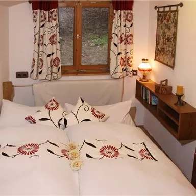 A cozy bedroom with a double bed and decorative pillows. The windows are adorned with red curtains and there is a small wooden shelf wall.