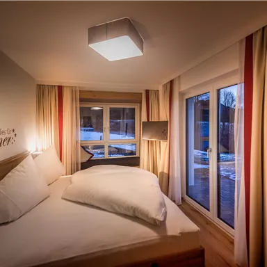 Ein gemütliches Schlafzimmer mit einem großen Bett und hellen Vorhängen. Große Fenster bieten einen Blick nach draußen.