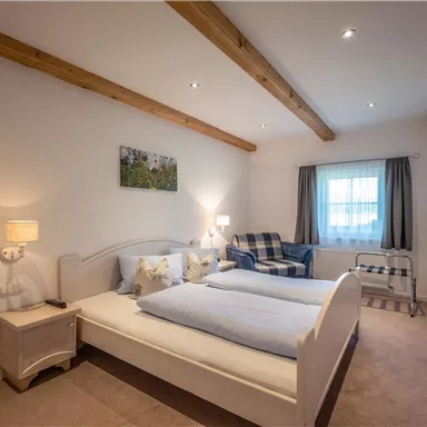 A cozy bedroom with a double bed and an additional bed. The room has wooden ceiling beams, a comfortable seating area, and a window with curtains.