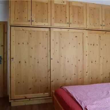 Ein einfaches Schlafzimmer mit einem großen, hölzernen Kleiderschrank und einem Bett in der Ecke. Die Wände sind hell und die Atmosphäre ist gemütlich.