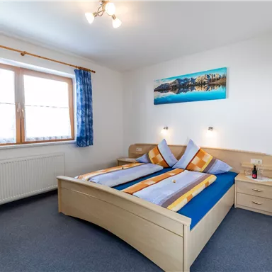 Ein helles Schlafzimmer mit einem Doppelbett und blauen Vorhängen. An der Wand hängt ein Bild einer Landschaft und es gibt eine kleine Lampe daneben.