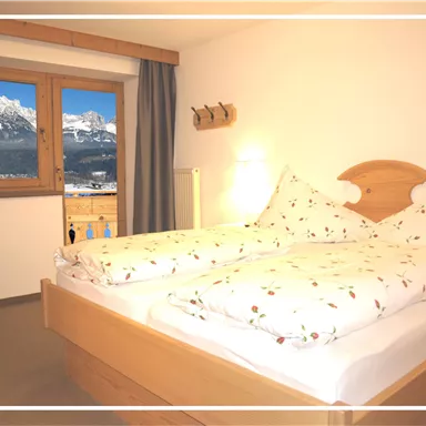 A cozy bedroom with two beds and floral bedding. The view from the window shows impressive mountains and a clear sight.