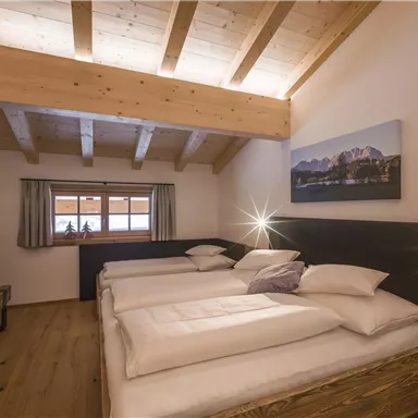 Ein helles und modernes Schlafzimmer mit Holzbalkendecke. Es gibt drei Betten, Fenster mit Vorhängen und ein schönes Landschaftsbild an der Wand.