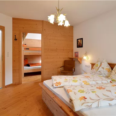 A cozy bedroom with wooden paneling and a large bed. In the background, there is an open room with bunk beds.