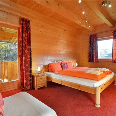 A cozy bedroom with wood paneling and a red carpet. The bed has orange bedding and large windows provide plenty of light.