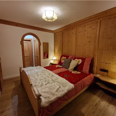 A cozy bedroom with a wooden wall design and a large bed. Notable details are the red cushions and the decorative lamps.