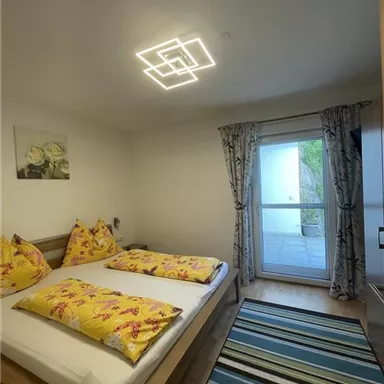 A bright bedroom with a double bed and colorful cushions. There is access to a balcony and a modern light shade.