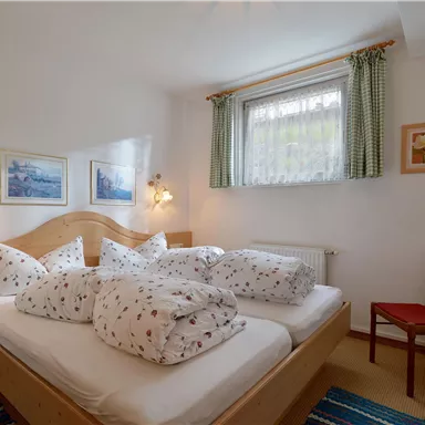 A cozy bedroom with a double bed and many cushions. There is a window with curtains and a chair in the corner.
