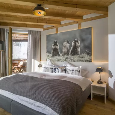 Ein modernes Schlafzimmer mit einem großen Bett und Holzdecke. Am Wand ist ein Bild von drei Tieren zu sehen, während viel Tageslicht durch das Fenster strömt.