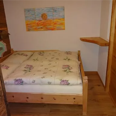 A cozy bedroom with a wooden bed and a mattress covered in a floral pattern. A colorful picture hangs on the wall, and there is a small side table.