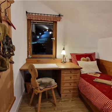 A cozy bedroom with a bed, a desk, and a wooden chair. It has a warm atmosphere with wooden paneling and a window that looks out onto the night.