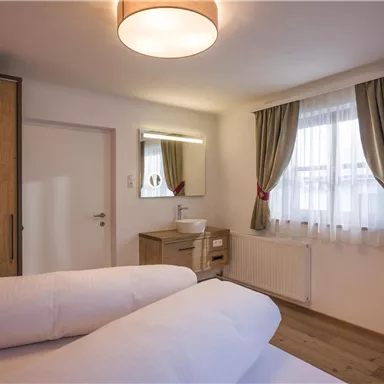 A cozy room with two beds, a wooden wardrobe, and a large window. The walls are light, and there is a modern television on the wall.