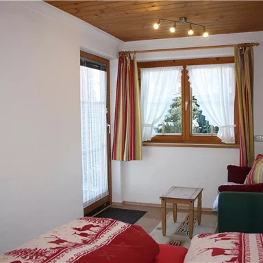 Ein gemütliches Zimmer mit einem Fenster, das viel Licht hereinlässt. Farbig gestreifte Vorhänge und eine kleine Sitzecke sorgen für eine einladende Atmosphäre.