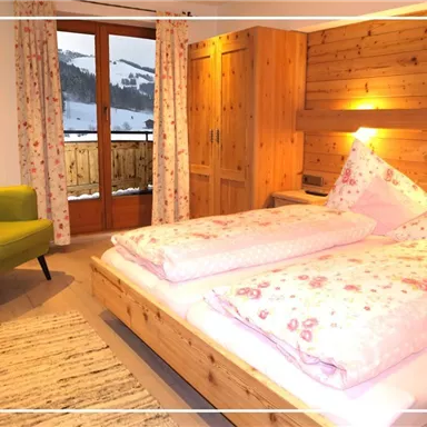 A cozy bedroom with a wooden interior and a large window overlooking a snowy landscape. There is a bed with floral comforters and a green armchair in the corner.
