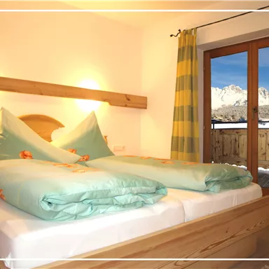 A cozy bedroom with wooden furniture and colorful bedding. The view shows snow-covered mountains through the window.