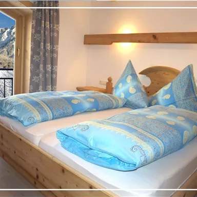 A cozy bedroom with a large bed and blue pillows. In the background, there are windows overlooking a mountain landscape.