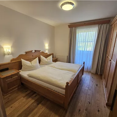 A cozy bedroom with two beds and wooden furniture. The room is bright and inviting, with a window that lets in daylight.