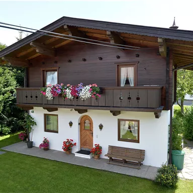 A cozy house with an upper balcony, decorated with colorful flowers. The garden is green and inviting, surrounded by trees.
