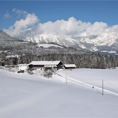 Eine verschneite Landschaft mit schneebedeckten Bergen im Hintergrund. Ein Bauernhaus und einige Wanderer sind im Vordergrund sichtbar.